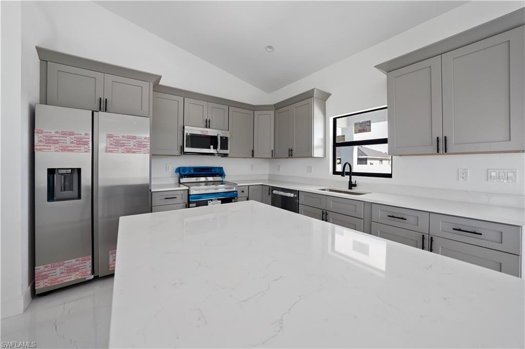 Kitchen with gray cabinets, stainless steel appliances, light stone counters, lofted ceiling, and light marble finish flooring