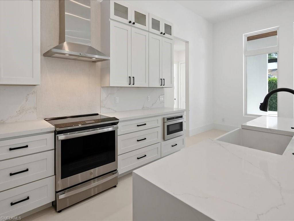 Kitchen with stainless steel electric stove, wall chimney range hood, light stone countertops, and white cabinetry