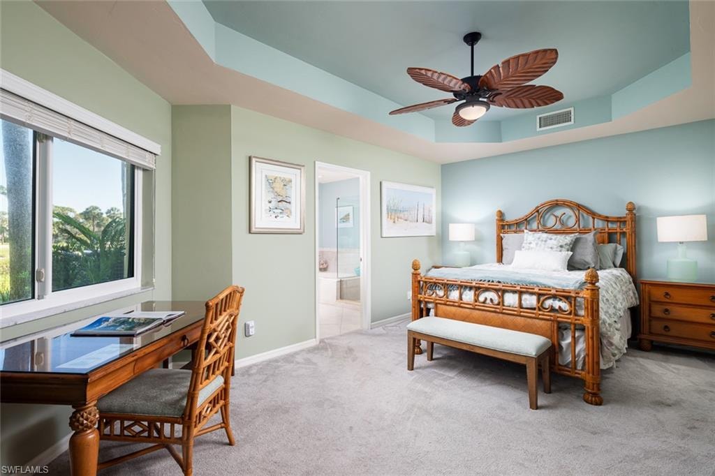 Carpeted bedroom with visible vents, a tray ceiling, baseboards, a ceiling fan, and ensuite bath
