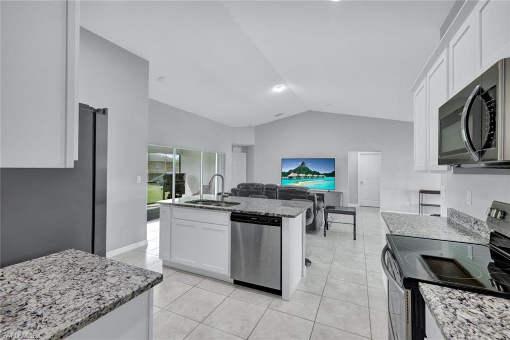 Kitchen featuring appliances with stainless steel finishes, white cabinets, lofted ceiling, a center island with sink, and light stone counters