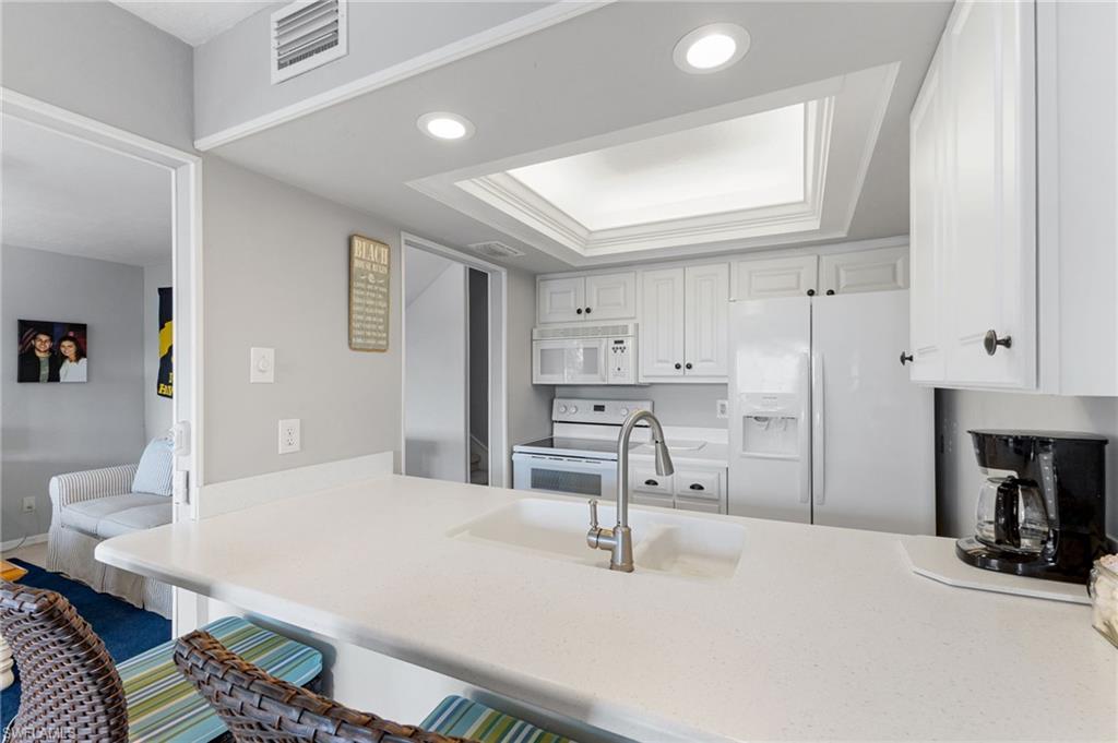 Kitchen with a raised ceiling, white cabinetry, white appliances, a kitchen breakfast bar, and recessed lighting
