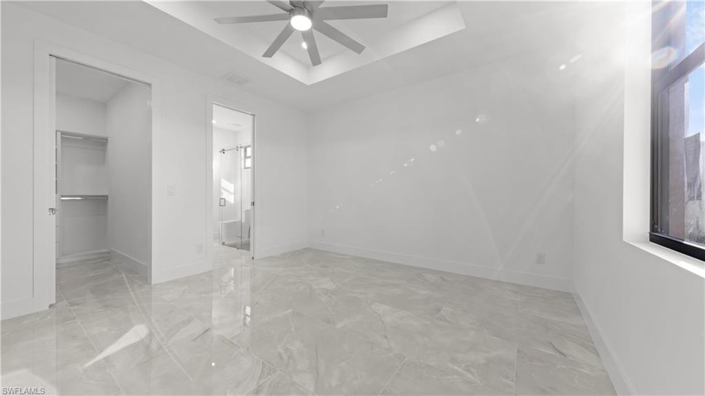 Unfurnished bedroom with a tray ceiling, a spacious closet, ceiling fan, light marble finish floors, and ensuite bathroom