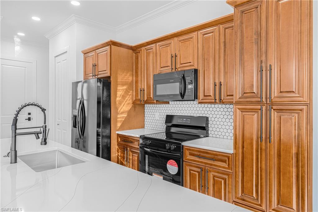 Kitchen with black appliances, ornamental molding, light stone counters, brown cabinetry, and decorative backsplash