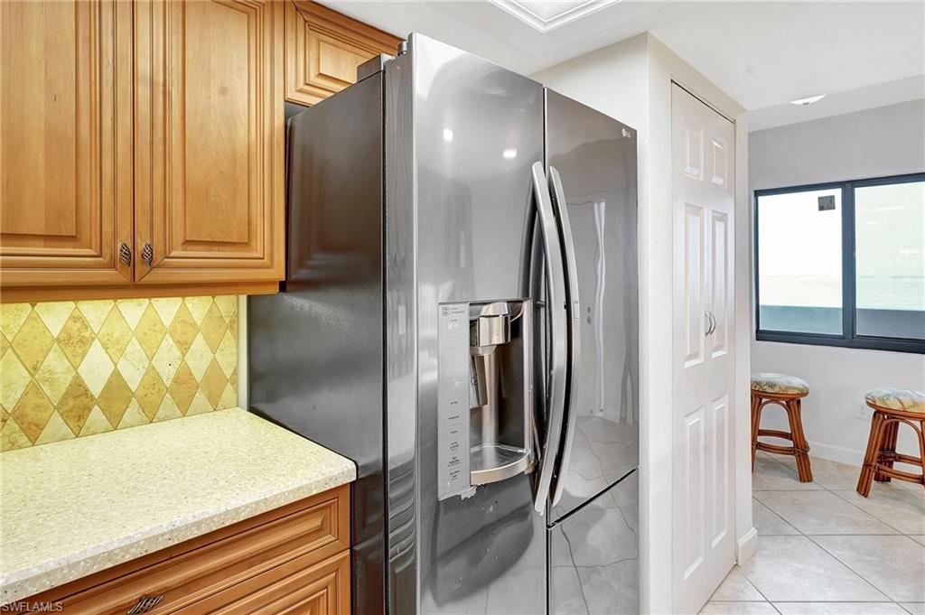 Kitchen featuring stainless steel fridge, light stone countertops, light tile patterned floors, brown cabinets, and tasteful backsplash