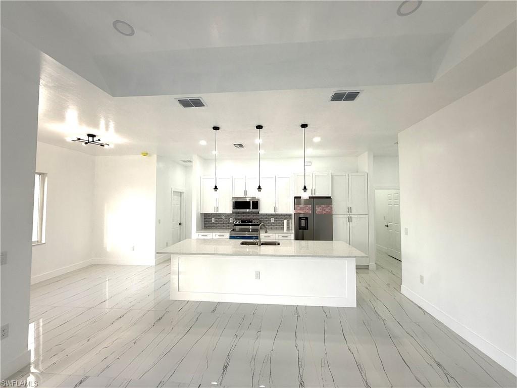 Kitchen featuring appliances with stainless steel finishes, white cabinets, tasteful backsplash, a kitchen island with sink, and light stone countertops