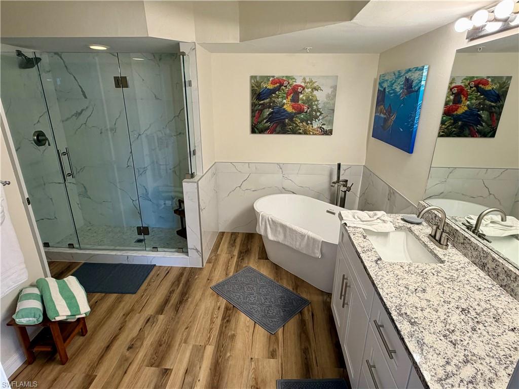 Modern bathroom featuring a frameless glass shower enclosure with white marbled tile surround, a freestanding soaking tub, a single vanity with a light-colored speckled countertop, wood-finish flooring, and a large wall-mounted mirror