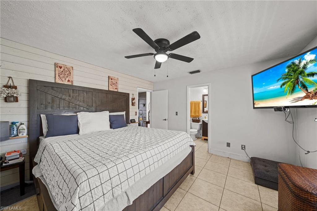 Bedroom with a textured ceiling, wood walls, a ceiling fan, light tile patterned floors, and connected bathroom