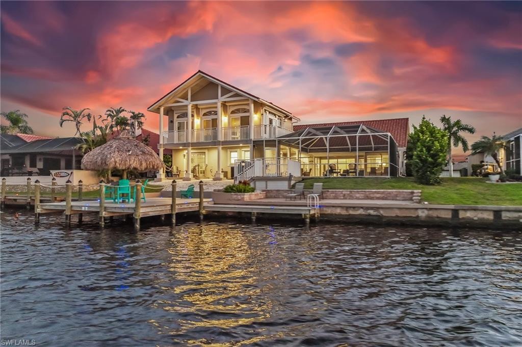 Back of house at dusk with a water view, a patio, glass enclosure, stucco siding, and a yard
