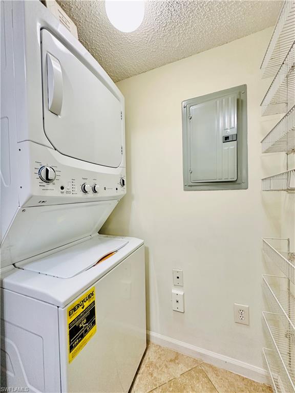 Washroom featuring electric panel, a textured ceiling, stacked washer / drying machine, and light tile patterned floors