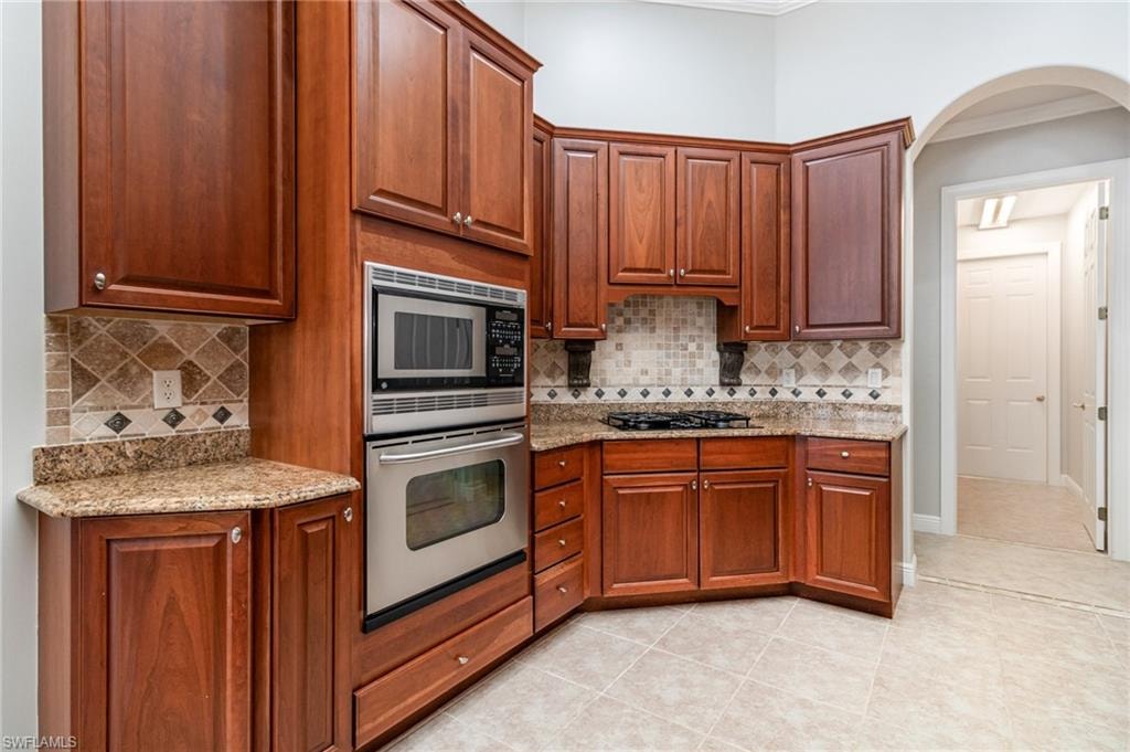 Kitchen with light stone countertops, stainless steel appliances, light tile patterned flooring, tasteful backsplash, and brown cabinets