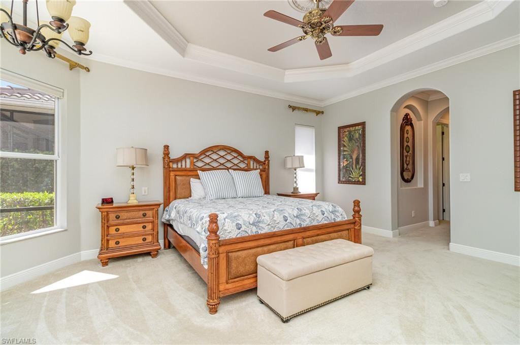 Carpeted bedroom with a raised ceiling, arched walkways, a ceiling fan, crown molding, and a chandelier