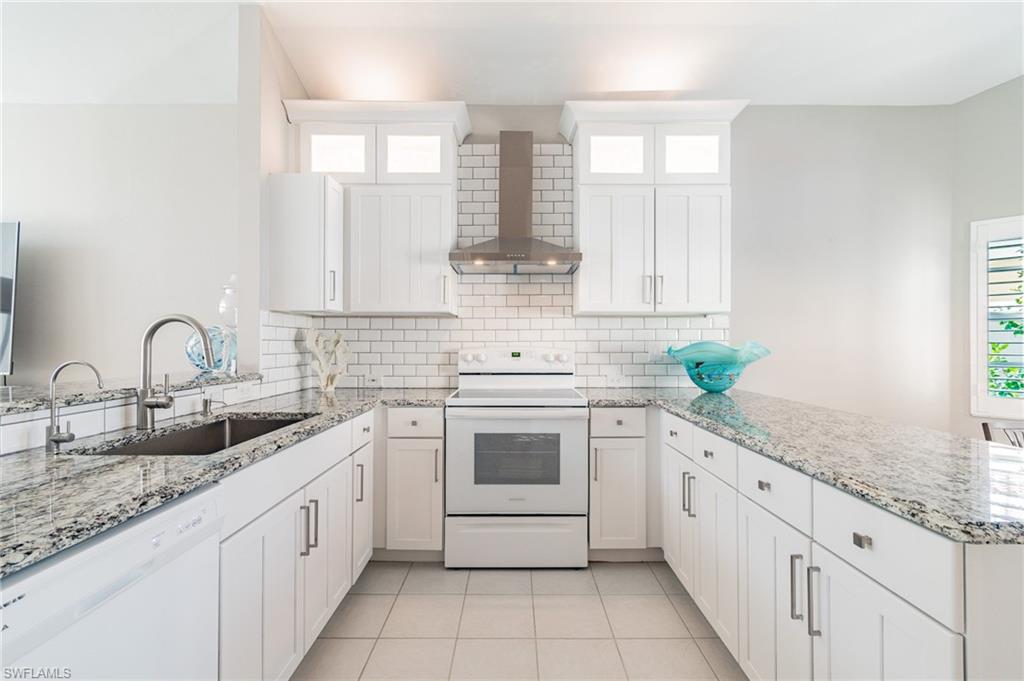Kitchen featuring a peninsula, white appliances, wall chimney range hood, light stone countertops, and white cabinets