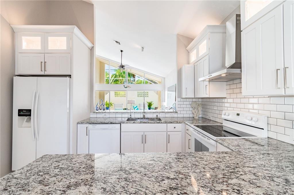 Kitchen featuring white appliances, glass insert cabinets, wall chimney exhaust hood, white cabinets, and vaulted ceiling