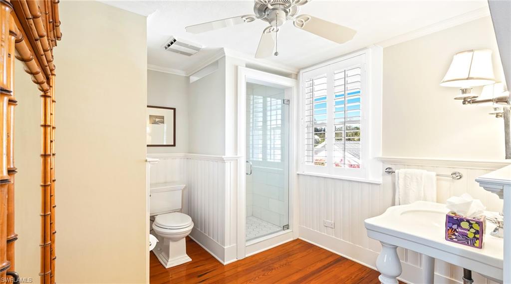 Full bath featuring crown molding, a shower stall, dark wood-type flooring, a wainscoted wall, and a ceiling fan