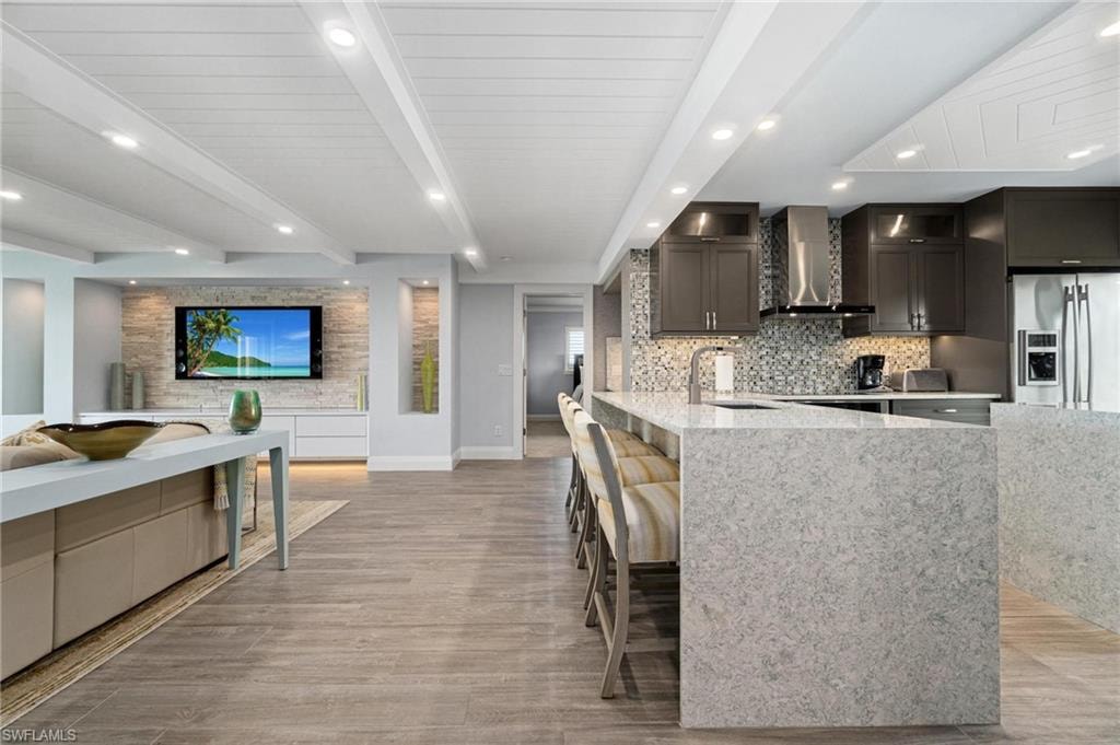 Kitchen with beam ceiling, stainless steel fridge with ice dispenser, a kitchen bar, wall chimney range hood, and light stone counters