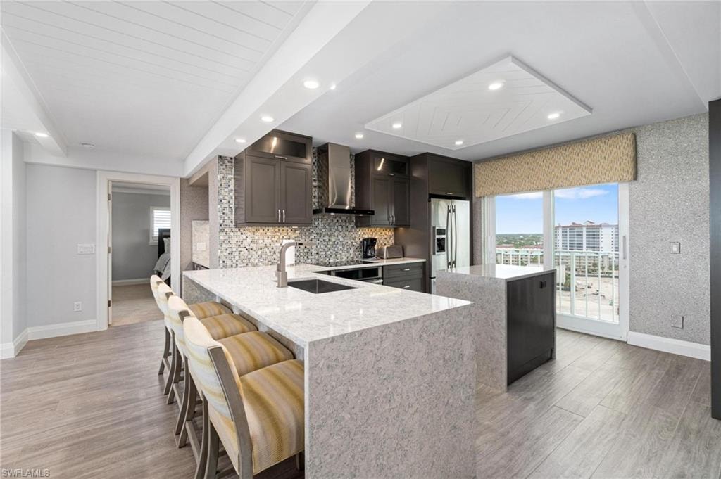 Kitchen featuring a peninsula, light stone counters, wall chimney exhaust hood, stainless steel refrigerator with ice dispenser, and a breakfast bar area