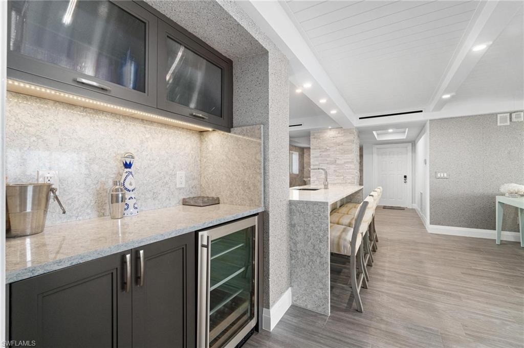 Bar featuring light stone counters, wine cooler, light wood-style floors, tasteful backsplash, and glass insert cabinets