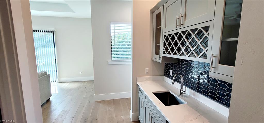 Upstairs wet bar with light wood finished floors, light stone countertops, backsplash, glass insert cabinets, and gray cabinetry