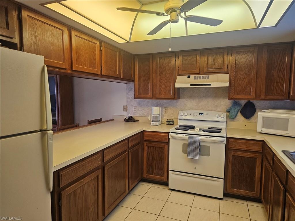 Kitchen with white appliances, light countertops, light tile patterned flooring, under cabinet range hood, and backsplash