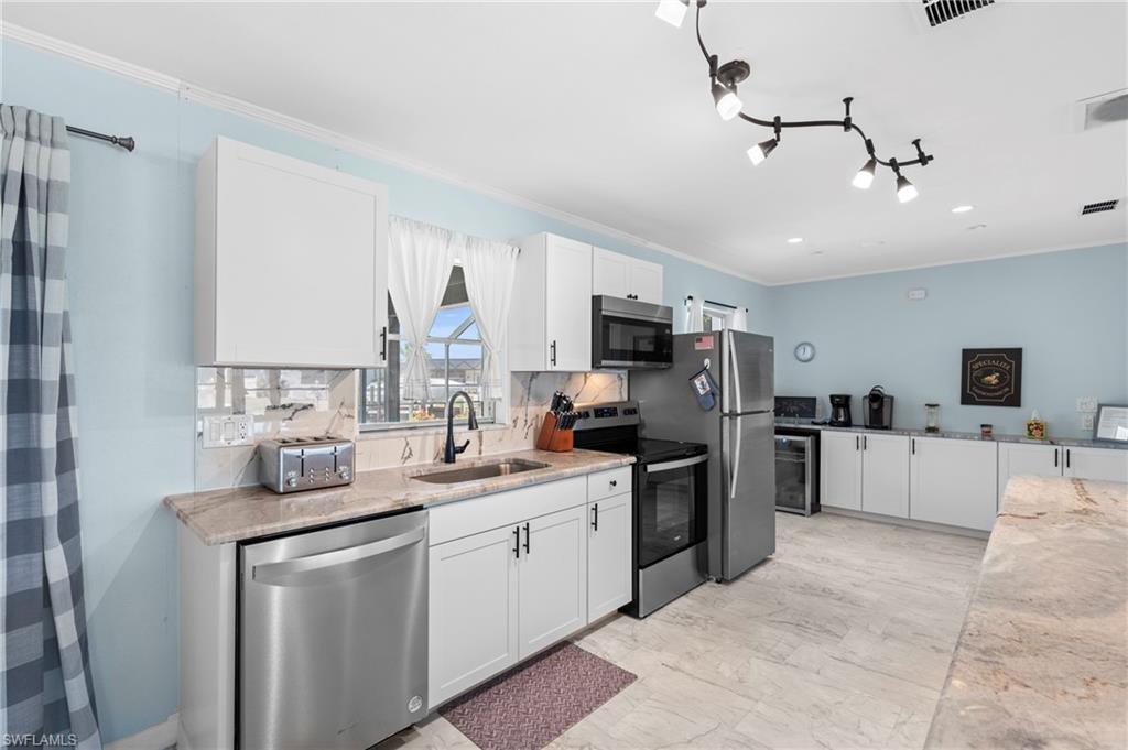 Kitchen with white cabinets, appliances with stainless steel finishes, light stone counters, ornamental molding, and wine cooler