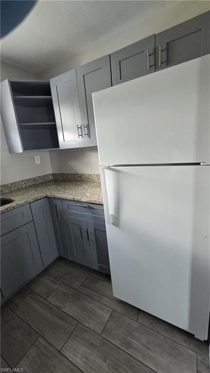 Kitchen featuring gray cabinets, freestanding refrigerator, light stone countertops, open shelves, and wood tiled floors