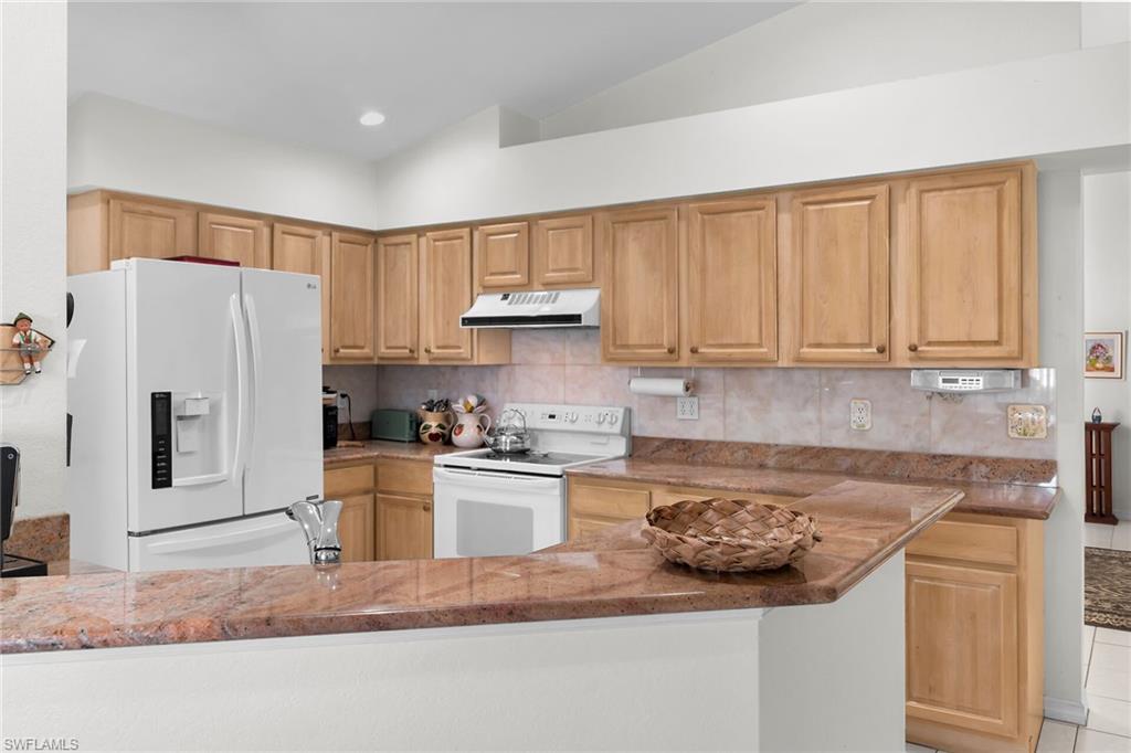 Kitchen with white appliances, tasteful backsplash, dark stone countertops, under cabinet range hood, and light brown cabinetry