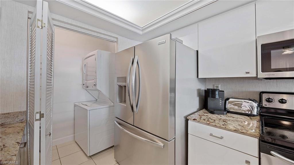 Kitchen with appliances with stainless steel finishes, white cabinetry, light stone countertops, light tile patterned flooring, and stacked washing machine and dryer