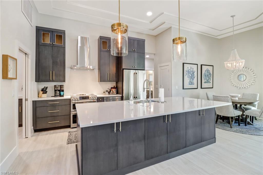 Kitchen with stainless steel appliances, wall chimney exhaust hood, decorative light fixtures, a large island with sink, and light stone counters