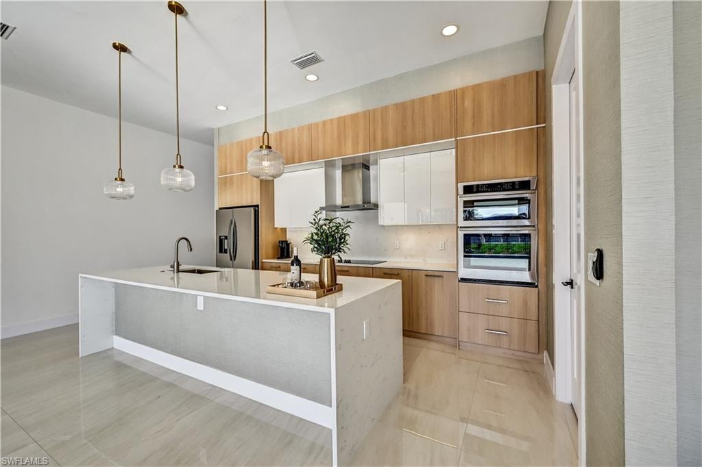Dual tone kitchen featuring decorative light fixtures, two tone color scheme, stainless steel appliances, a center island with sink, and light stone counters