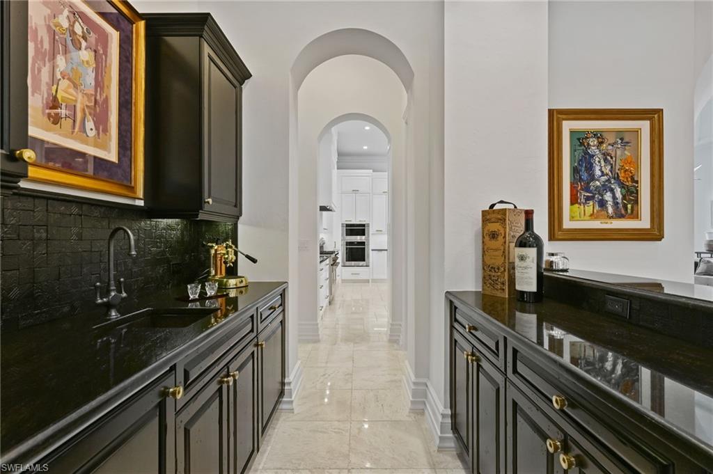 Indoor bar featuring arched walkways, dark stone counters, light marble finish flooring, decorative backsplash, and dark cabinets