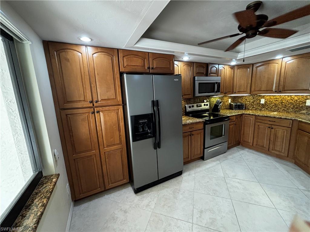 Kitchen with brown cabinets, appliances with stainless steel finishes, a raised ceiling, dark stone counters, and a ceiling fan