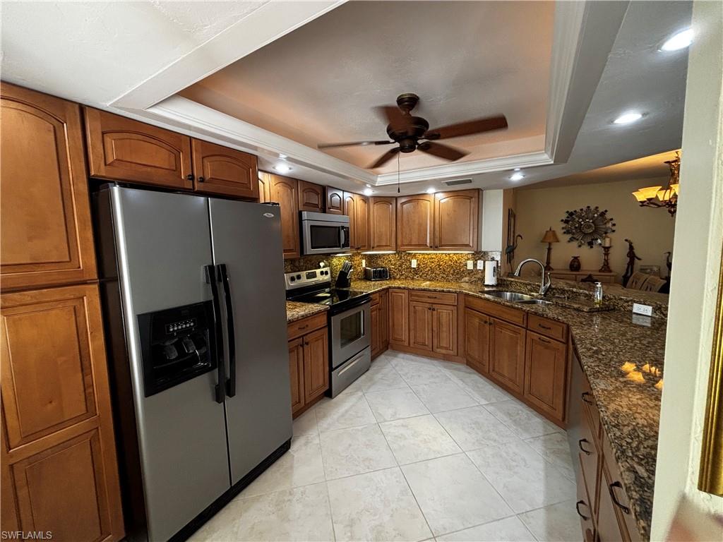 Kitchen with a tray ceiling, brown cabinetry, dark stone countertops, stainless steel appliances, and a chandelier