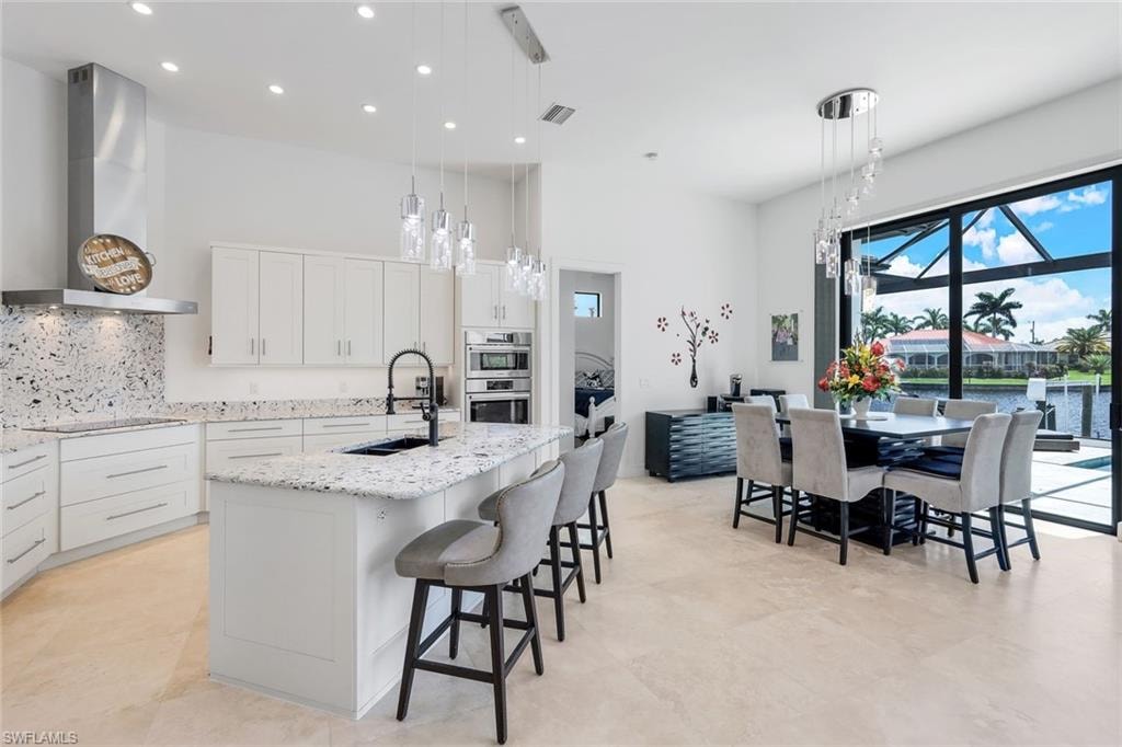 Kitchen featuring white cabinets, tasteful backsplash, pendant lighting, wall chimney range hood, and an island with sink