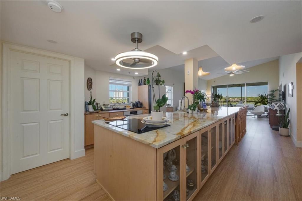 Kitchen with light stone counters, light wood-type flooring, glass insert cabinets, a ceiling fan, and a center island with sink