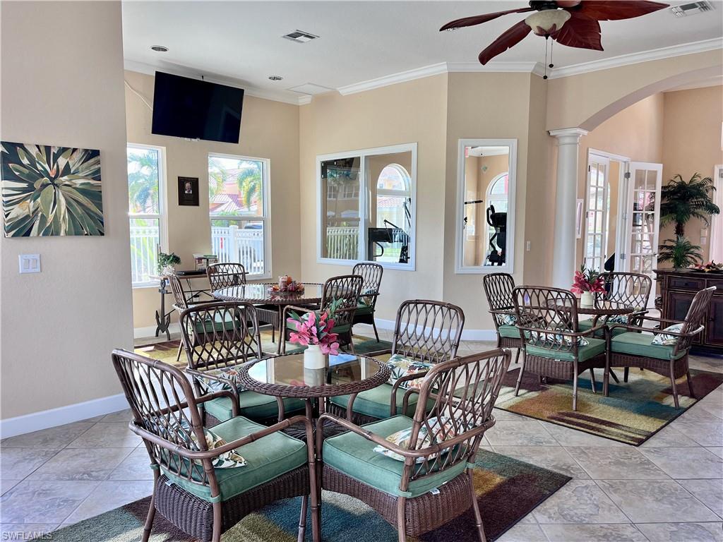 Community club house entertaining area with kitchen, tables and ornate columns, crown molding, light tile patterned flooring, arched walkways, and ceiling fan
