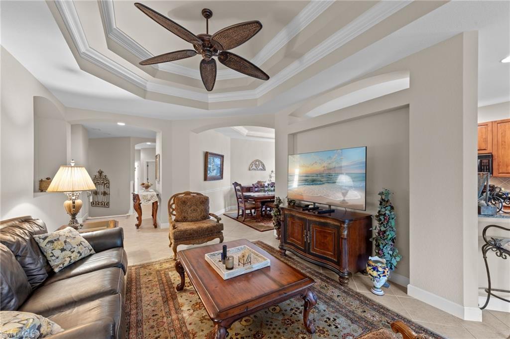 Living room with a tray ceiling, light tile patterned floors, arched walkways, ornamental molding, and ceiling fan