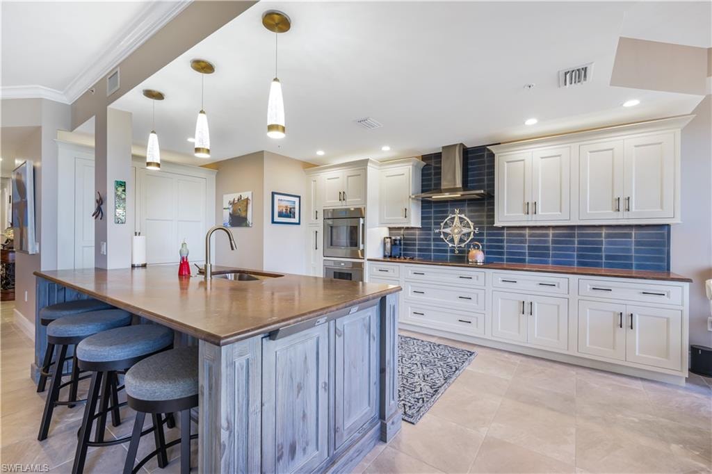Kitchen featuring tasteful backsplash, wall chimney range hood, a kitchen bar, white cabinets, and hanging light fixtures