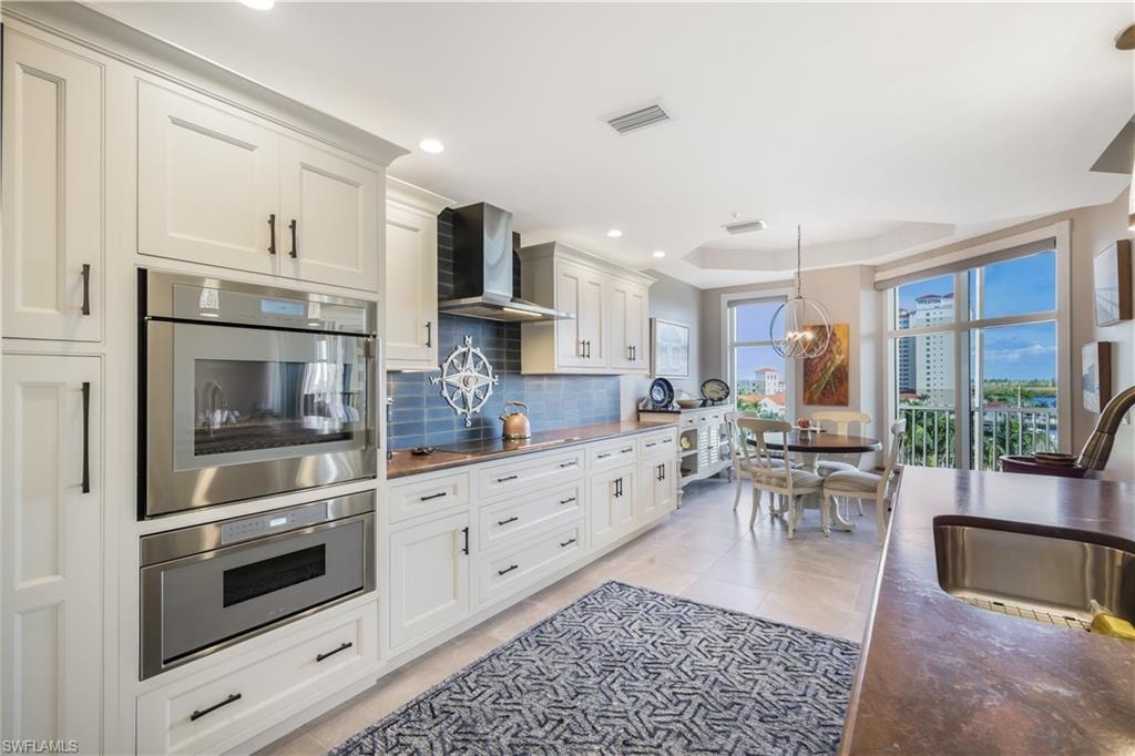 Kitchen featuring wall chimney range hood, stainless steel double oven, backsplash, decorative light fixtures, and white cabinetry