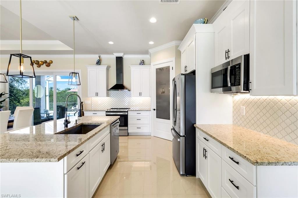Kitchen featuring light stone countertops, stainless steel appliances, white cabinetry, wall chimney range hood, and ornamental molding