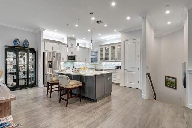 Featuring a large island with seating, hanging pending light fixtures, stainless steel appliances, and cabs with glass fronts and open staircase