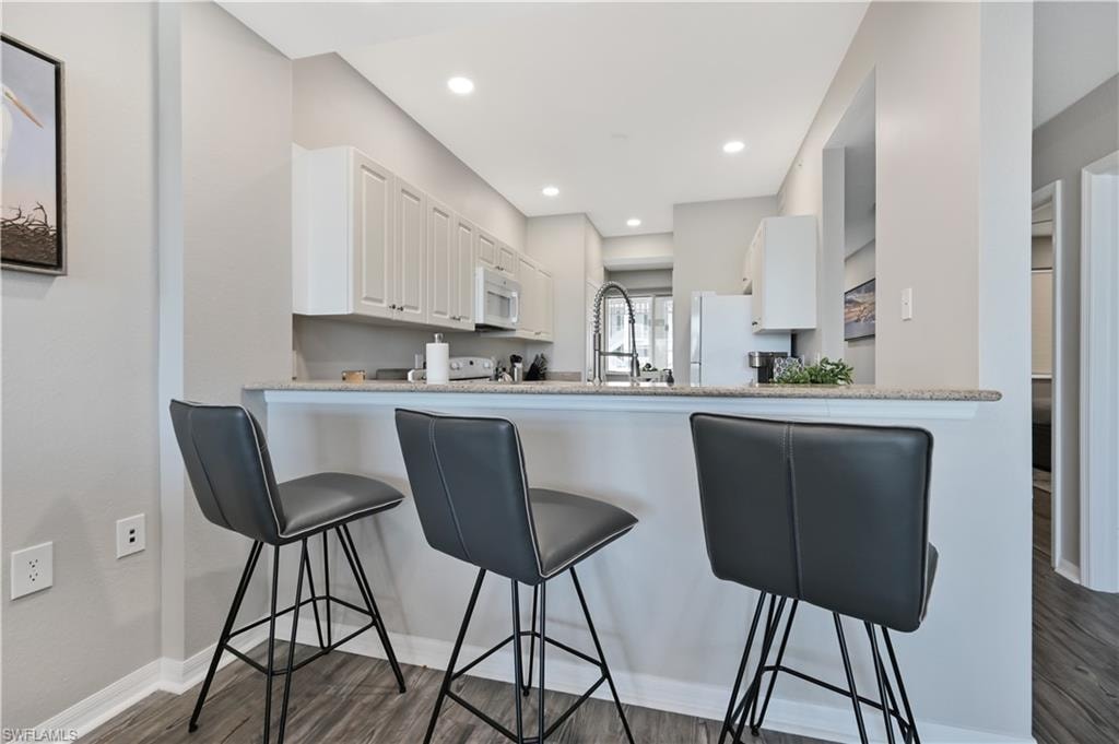 Kitchen with white cabinets, a breakfast bar, light stone counters, white appliances, and dark wood-type flooring