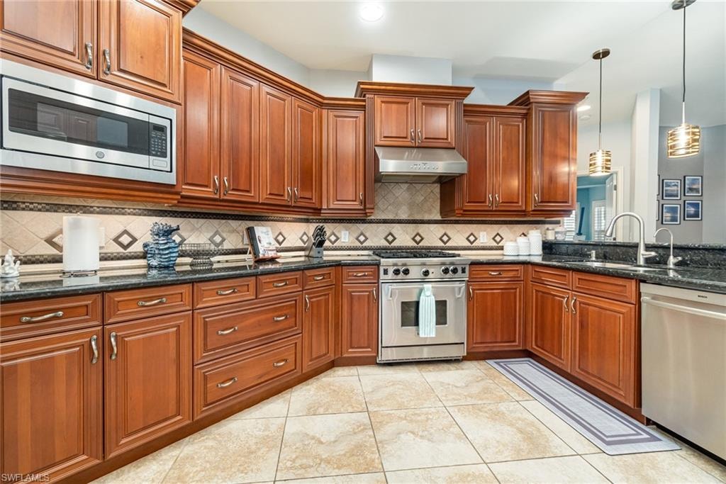 Kitchen featuring stainless steel appliances, hanging light fixtures, dark stone countertops, under cabinet range hood, and light tile patterned floors