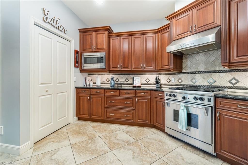 Kitchen featuring stainless steel appliances, under cabinet range hood, light tile patterned floors, dark stone counters, and backsplash
