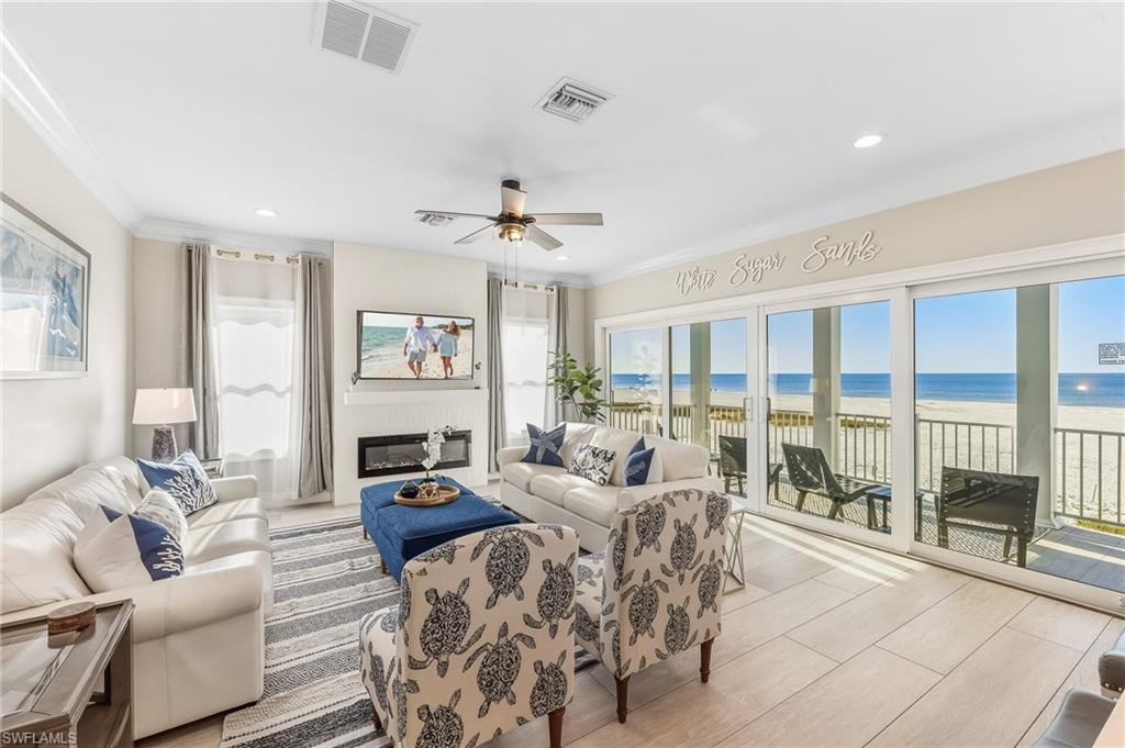 Living area with a ceiling fan, wood finished floors, view of water and beach, a glass covered fireplace, and ornamental molding