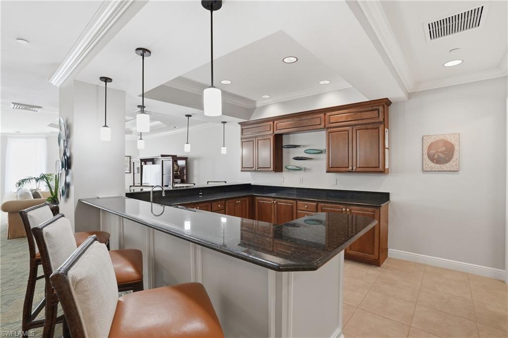 Community Kitchen featuring dark stone counters, ornamental molding, pendant lighting, a peninsula, and brown cabinetry - Virtually Edited Image