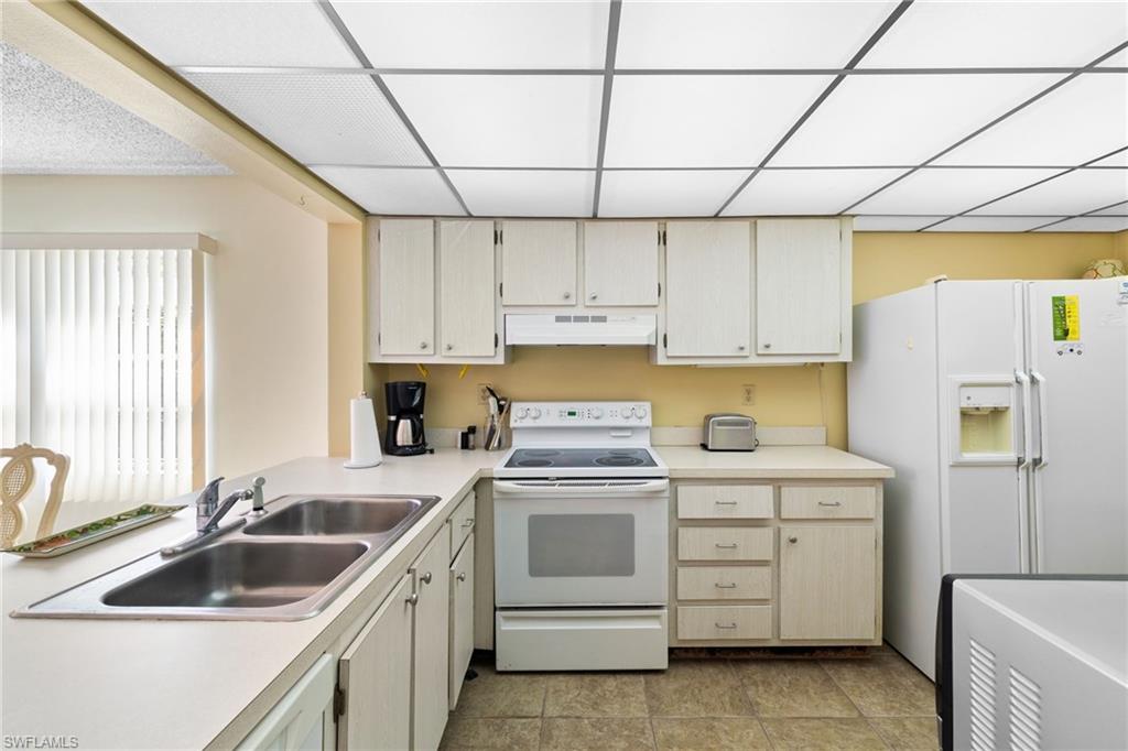 Kitchen with white appliances, light countertops, under cabinet range hood, a drop ceiling, and light tile patterned floors