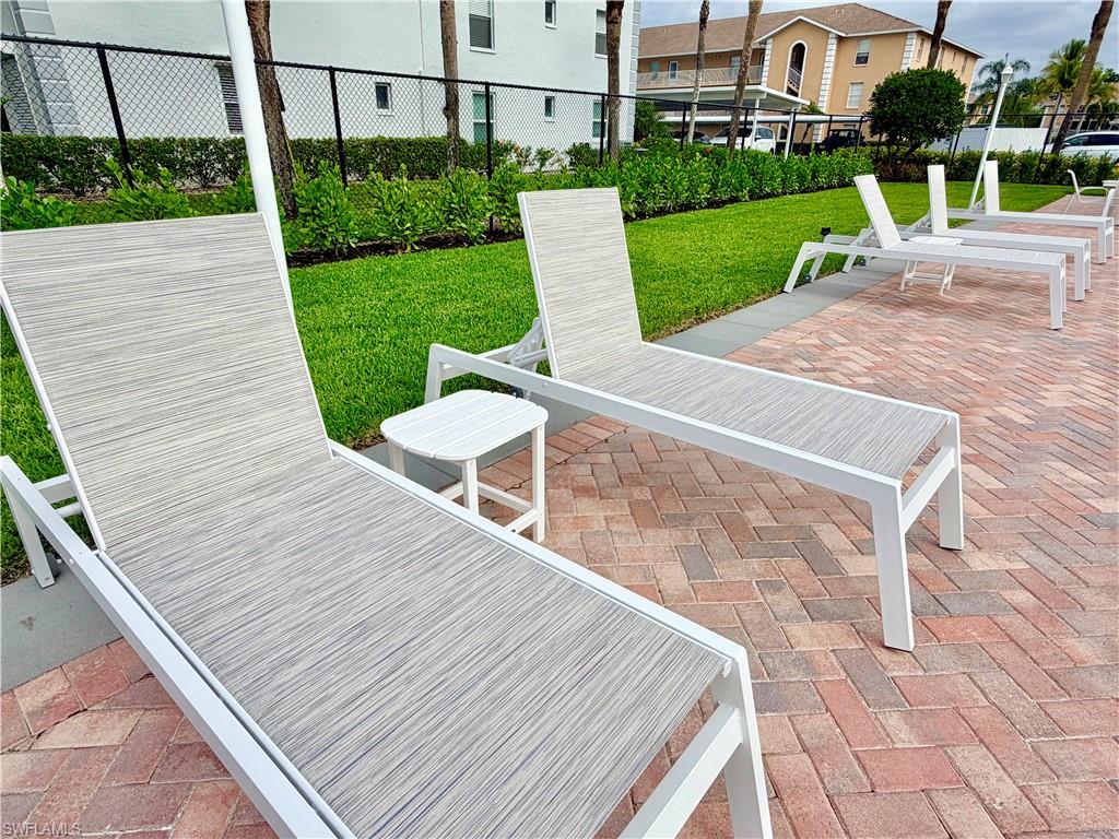 Outdoor lounge area featuring brick paver flooring, multiple lounge chairs with white frames, a white side table, manicured green lawn, and shrubbery