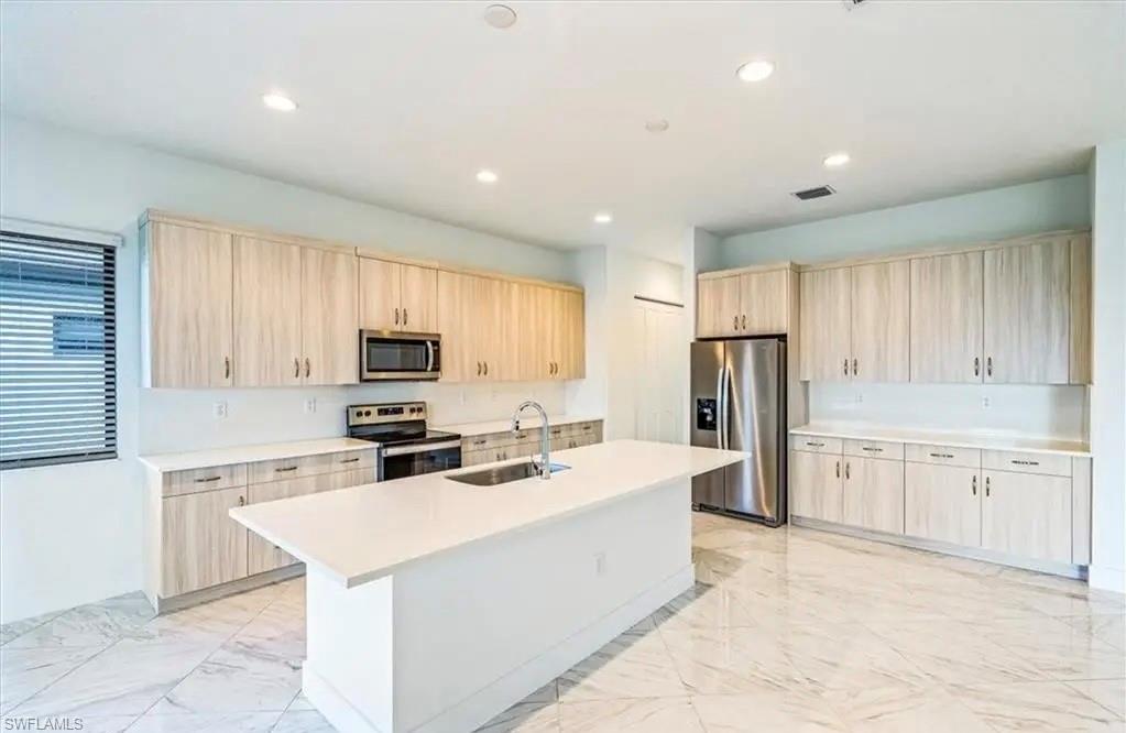 Kitchen featuring light brown cabinets, stainless steel appliances, light marble finish flooring, a kitchen island with sink, and recessed lighting
