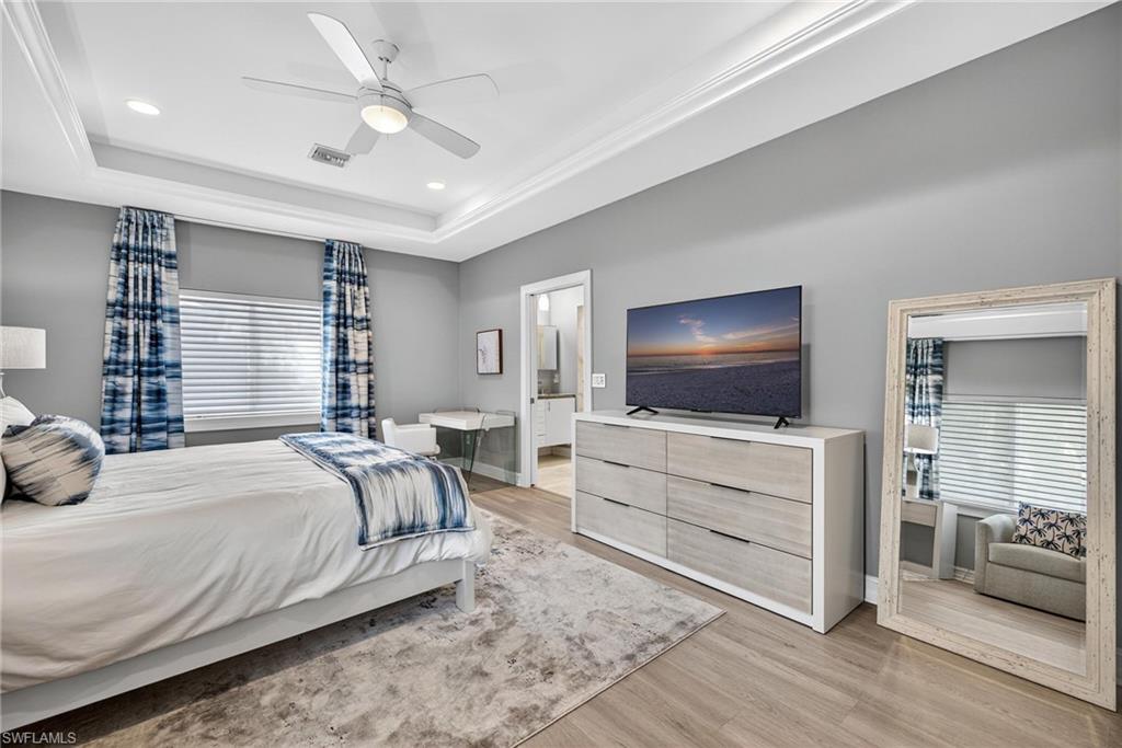 Bedroom with light wood-type flooring, ceiling fan, recessed lighting, connected bathroom, and a tray ceiling