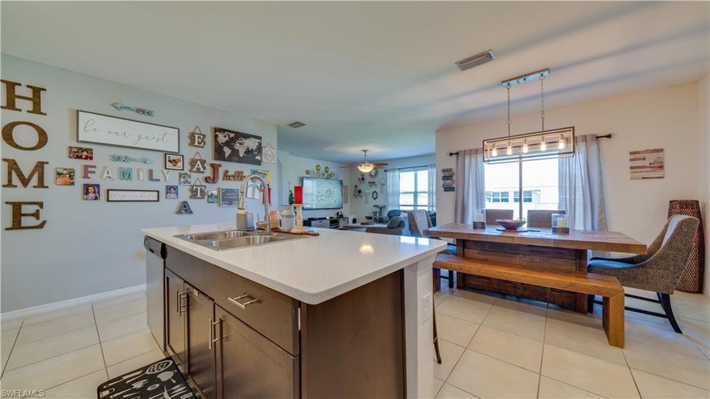 Kitchen featuring dark brown cabinets, open floor plan, an island with sink, light tile patterned flooring, and decorative light fixtures